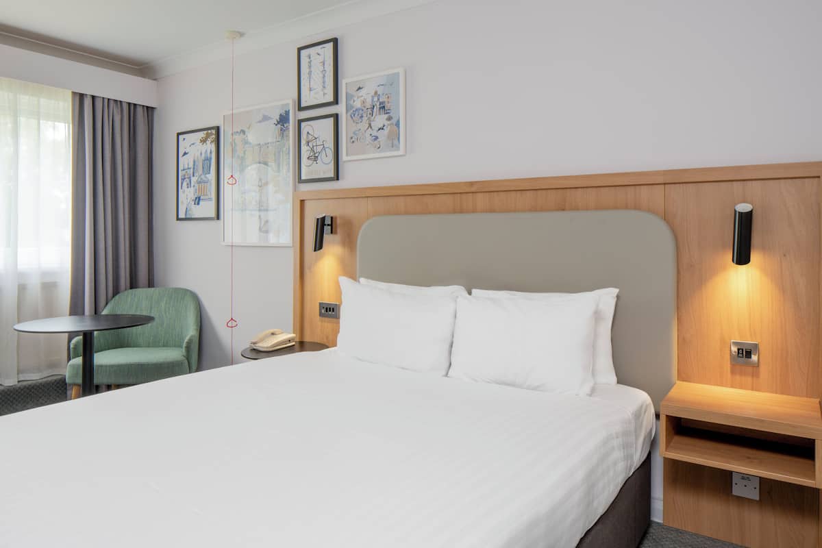 A hotel room with a large bed, a green armchair, a small round table, and artwork on the wall.