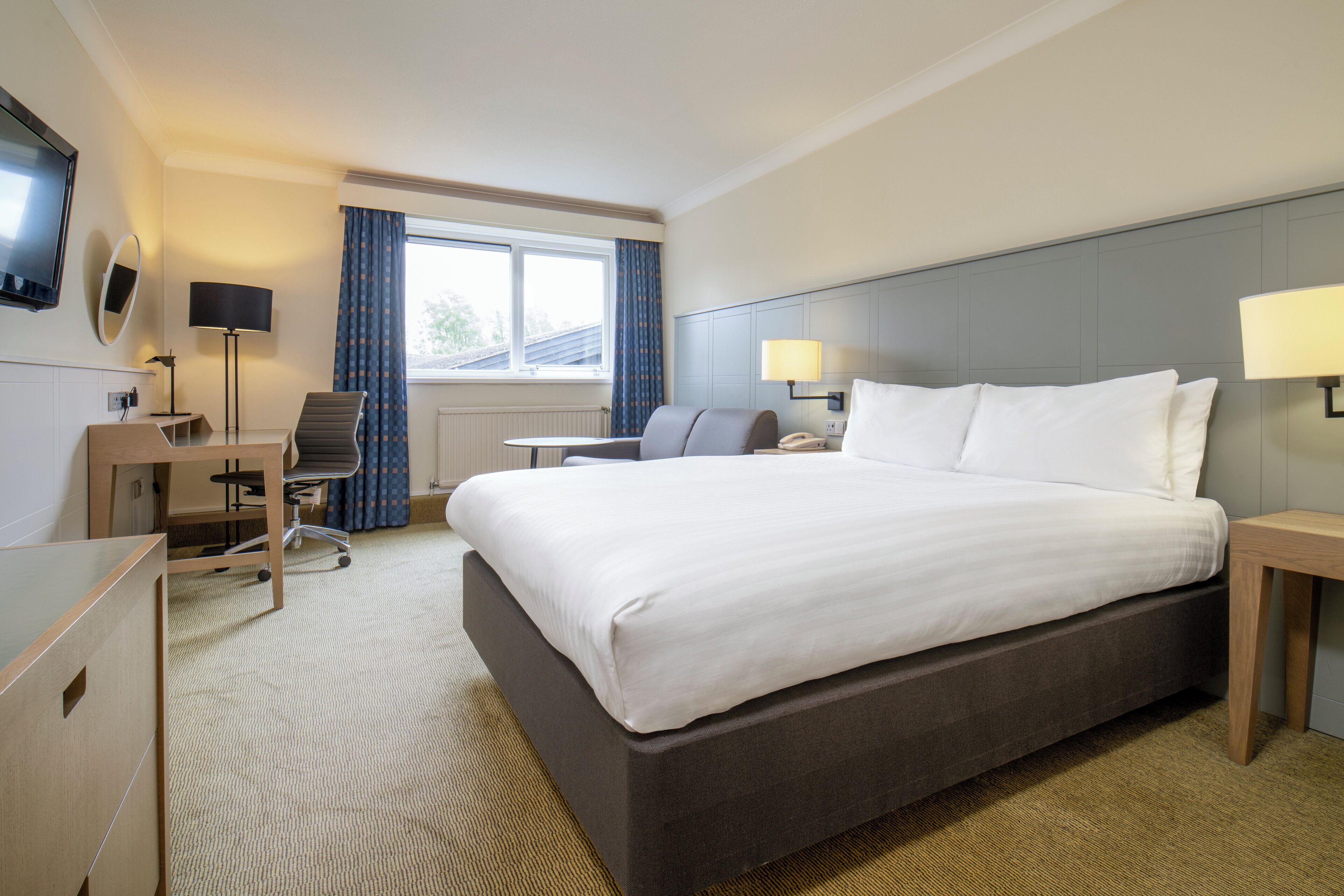 Une chambre d’hôtel avec un grand lit, un bureau, une chaise, une télévision et une fenêtre avec des rideaux.