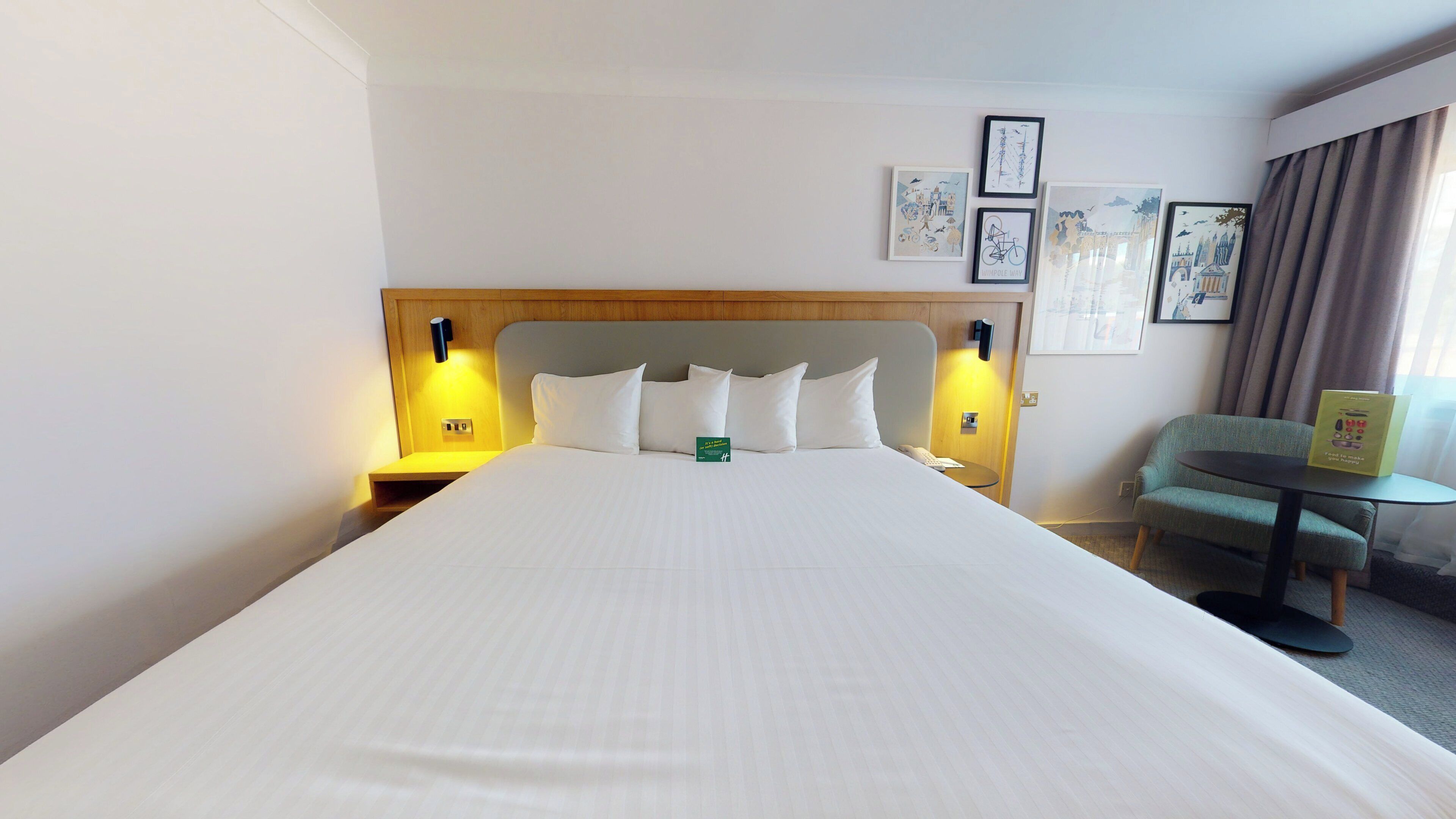 A hotel room with a large bed, two bedside tables with lamps, a small round table, a chair, and framed artwork on the wall.