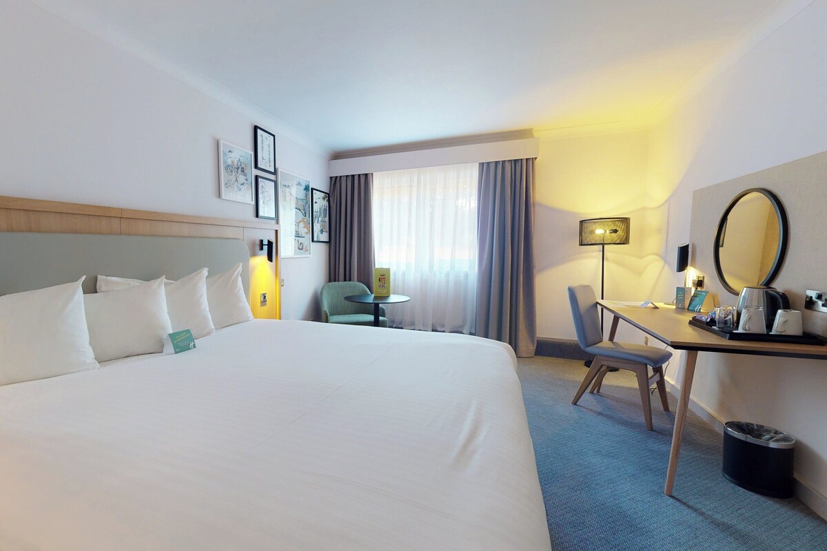 A hotel room with a large bed, a desk with a chair, a desk lamp, a trash can, and a mirror.
