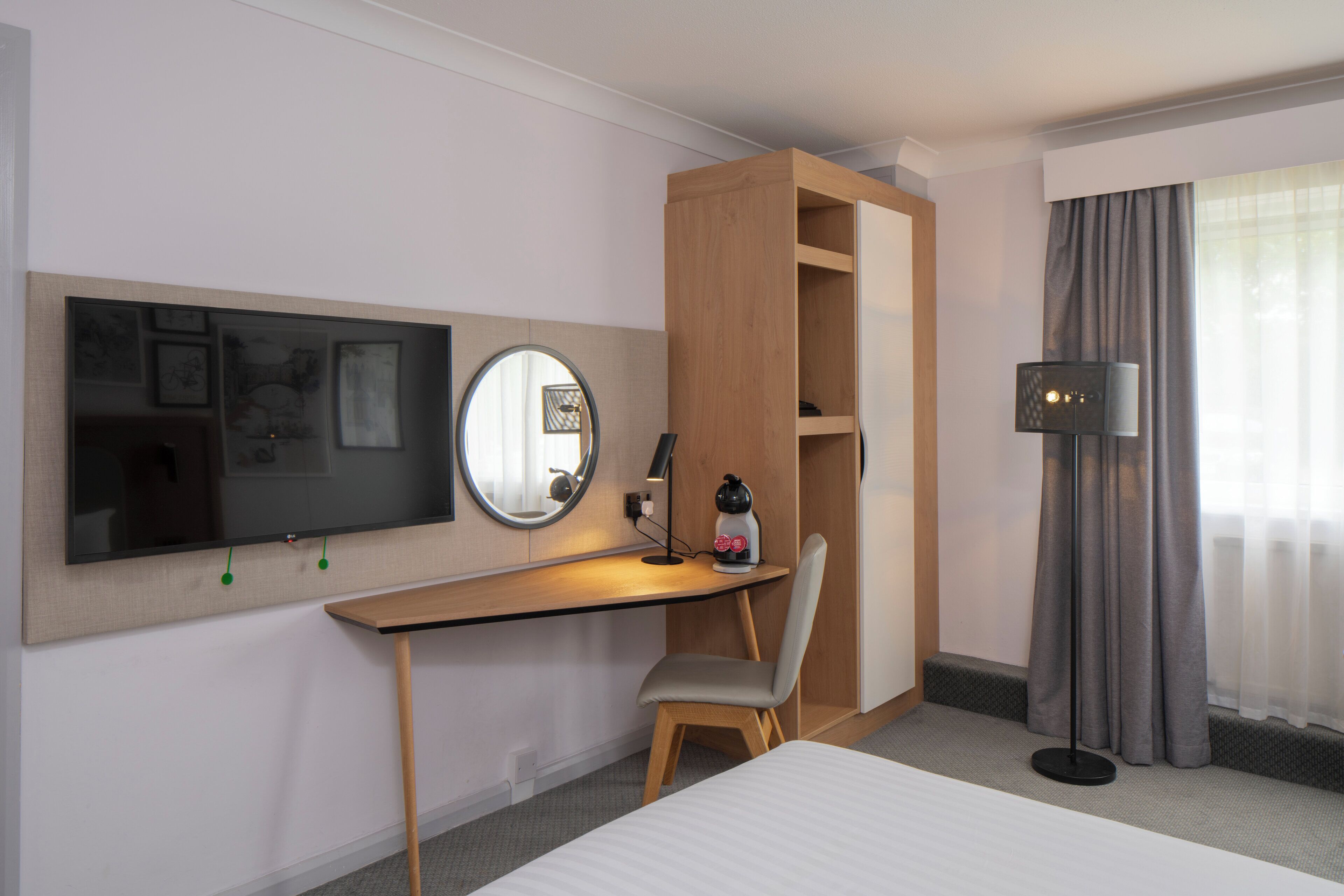 Habitación de hotel con cama, escritorio con silla, televisor, espejo, lámpara y armario.