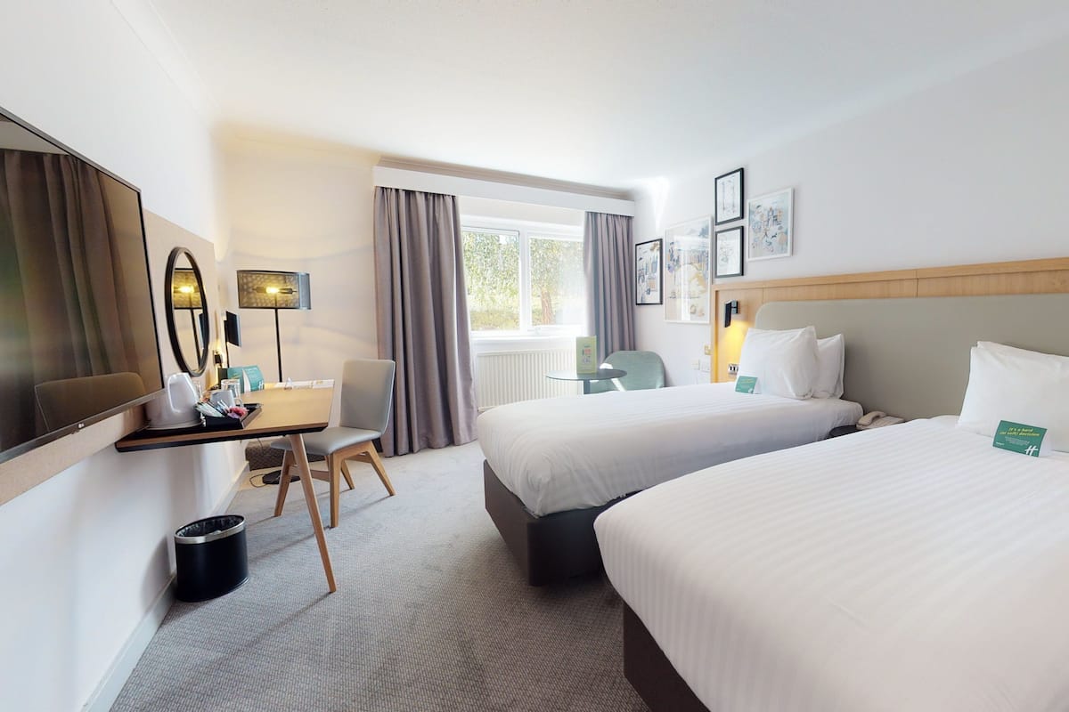 A hotel room with two beds, a desk with a chair, a mirror, and a window with curtains.