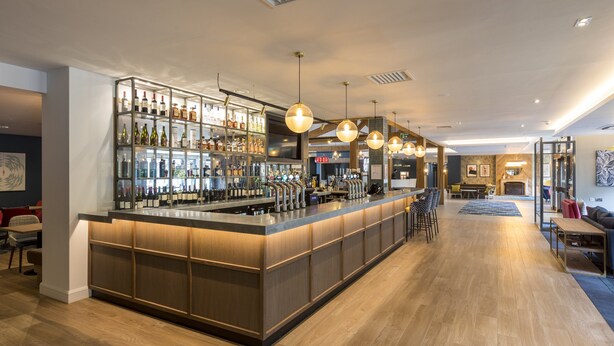 A modern bar area with a long counter, shelves of bottles, and hanging pendant lights. - Holiday Inn Cambridge by IHG (Cambridge)