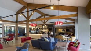 A spacious indoor area with wooden beams, a blue sofa, red chairs, and a bar area in the background. - Holiday Inn Cambridge by IHG (Cambridge)