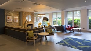 A modern lobby with a reception desk, seating area, and large windows offering a view of greenery. - Holiday Inn Cambridge by IHG (Cambridge)