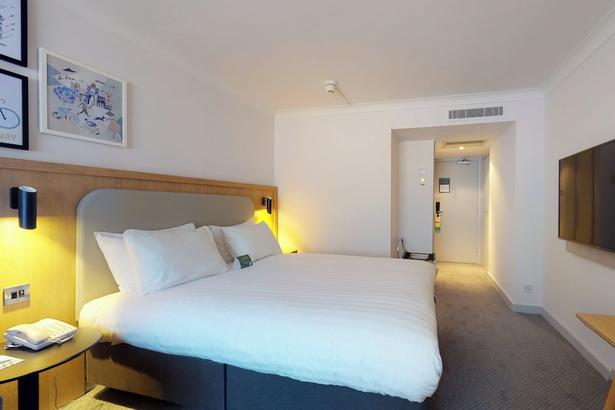 A hotel room with a large bed, a bedside table with a phone, a flat-screen TV mounted on the wall, and a hallway leading to another room.