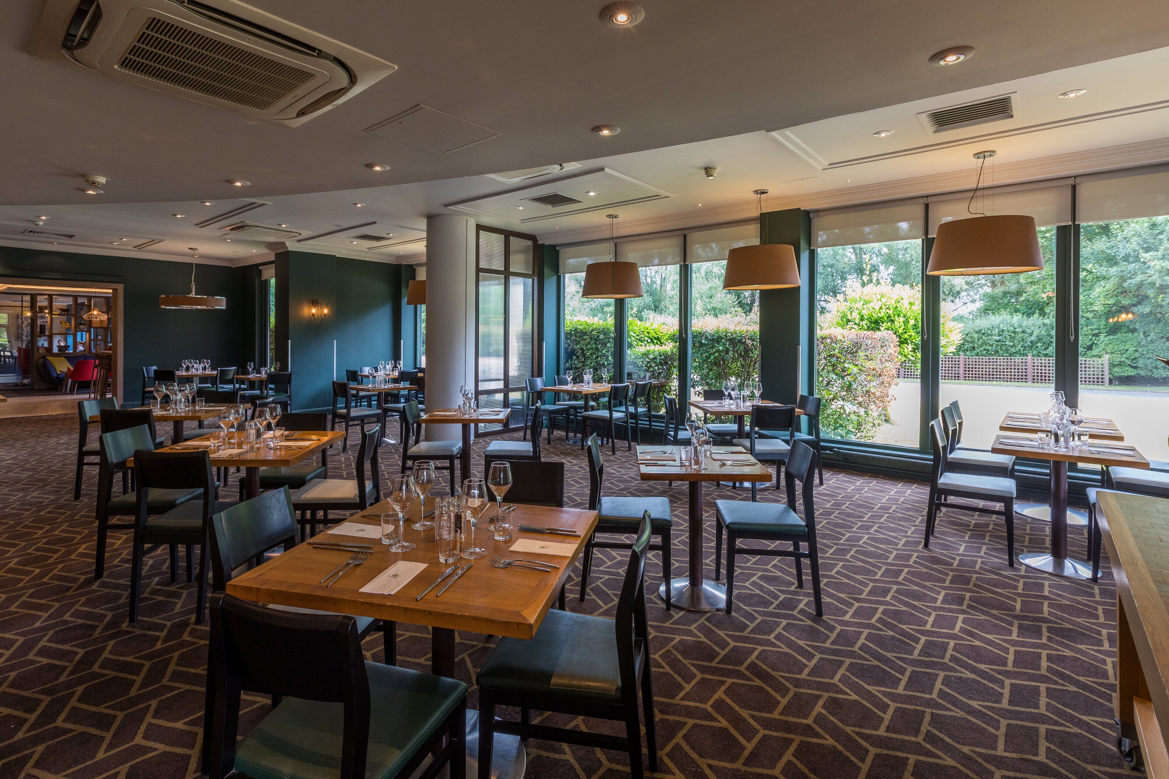 Un restaurante con mesas dispuestas para comer, grandes ventanales que dan a zonas verdes y una alfombra estampada.