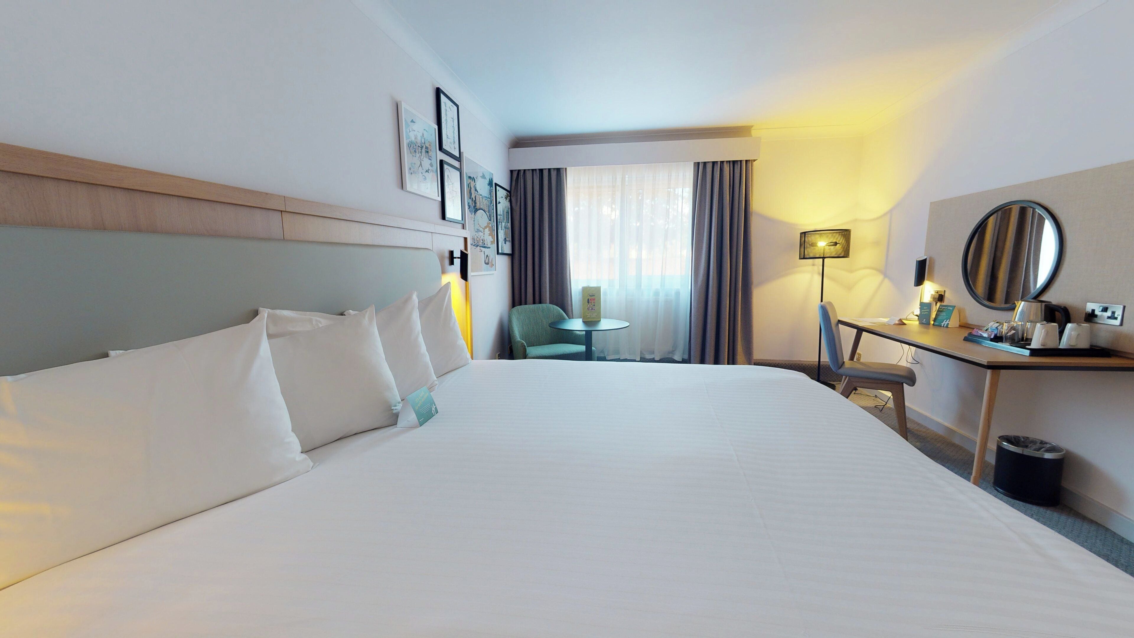 A hotel room with a large bed, a desk with a lamp, a chair, and a small table.