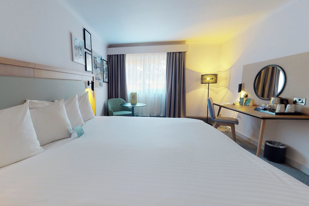 A hotel room with a large bed, a desk with a lamp, a chair, and a small table.
