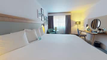 A hotel room with a large bed, a desk with a lamp, a chair, and a small table.
