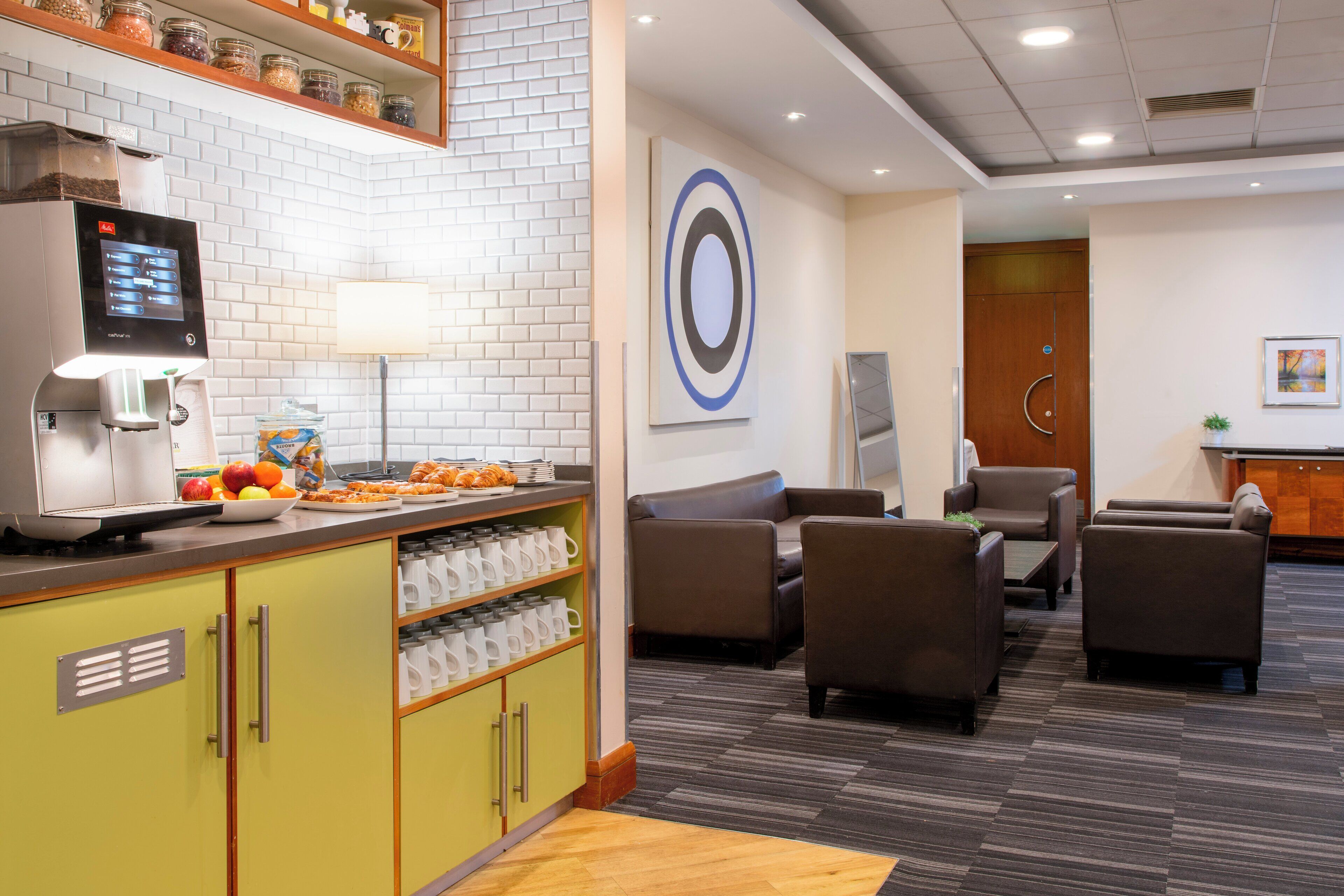 A modern hotel lobby with a coffee station, seating area, and a large wall art piece.