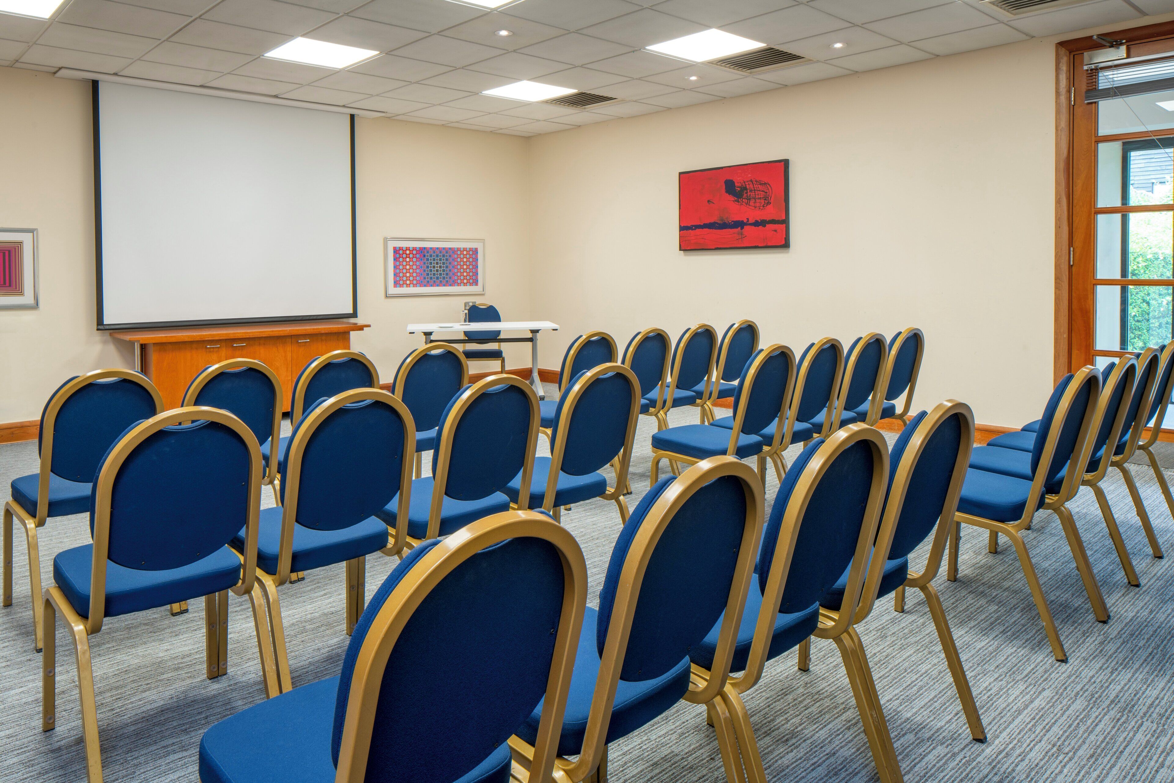 A conference room with rows of blue chairs, a podium, a projector screen, and a framed abstract painting on the wall.