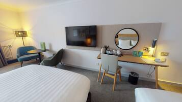 Una habitación de hotel con cama, escritorio con silla, televisor, espejo y una mesita de noche con lámpara.