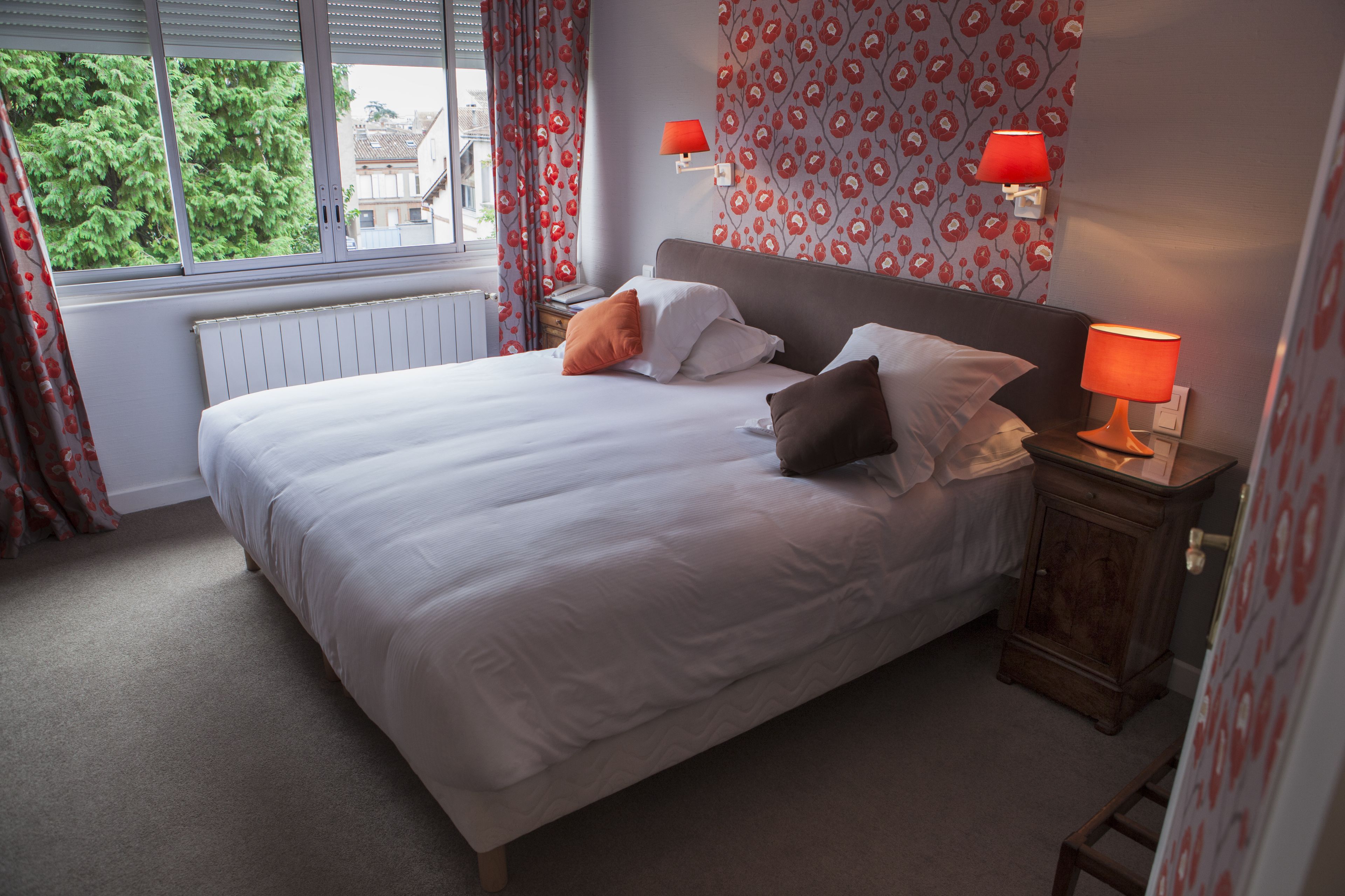 A neatly made bed with white linens and multiple pillows, a bedside table with a lamp, and a window with curtains.