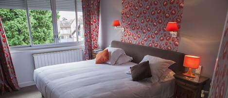 A neatly made bed with white linens and multiple pillows, a bedside table with a lamp, and a window with curtains.