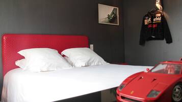 Une chambre d’hôtel avec un lit rouge, des oreillers blancs, une voiture de sport rouge et une veste accrochée au mur.