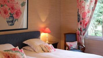 A hotel room with a bed, pillows, a nightstand with a lamp, a chair, and a large floral painting on the wall.