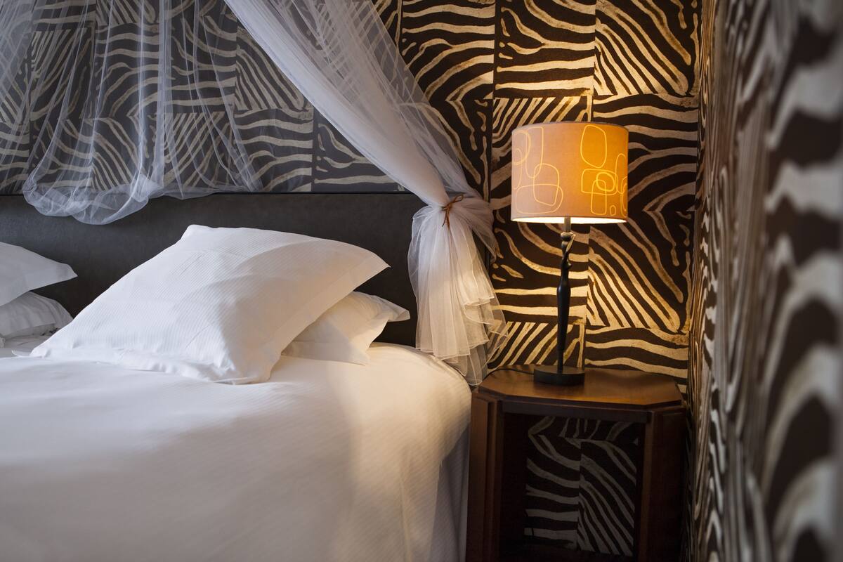 A neatly made bed with white linens, a bedside lamp with a zebra-patterned wall behind it, and sheer curtains over the bed.