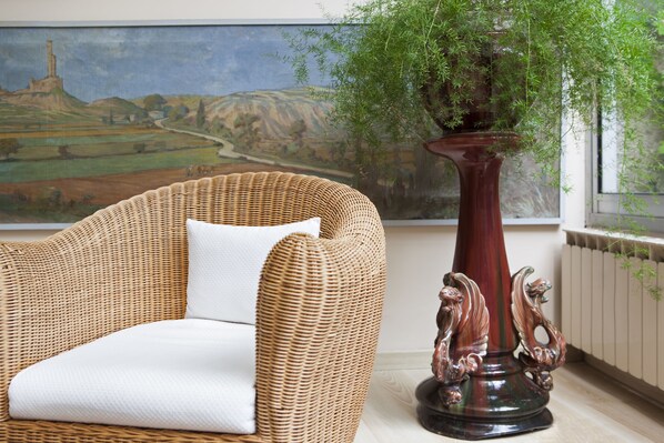 Een rieten bank met een witte kussen, een decoratieve vaas met een plant, en een schilderij van een landschap op de achtergrond.