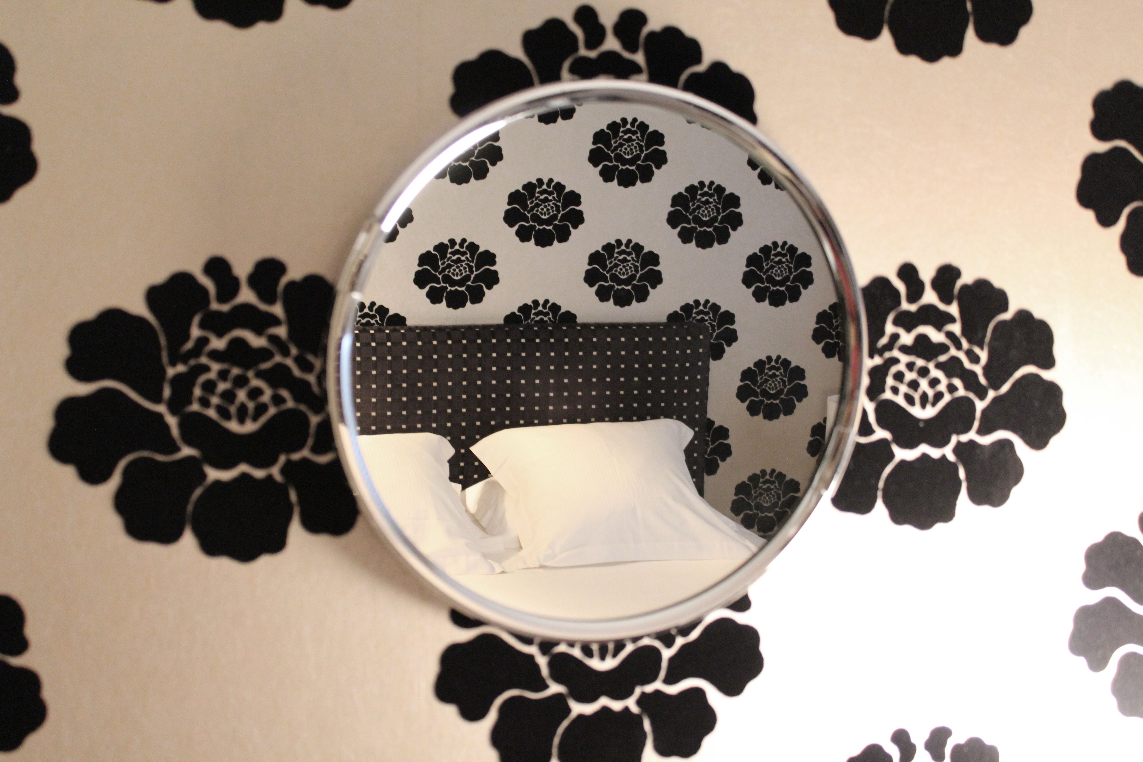A circular mirror with a floral pattern, reflecting a bed with white linens.