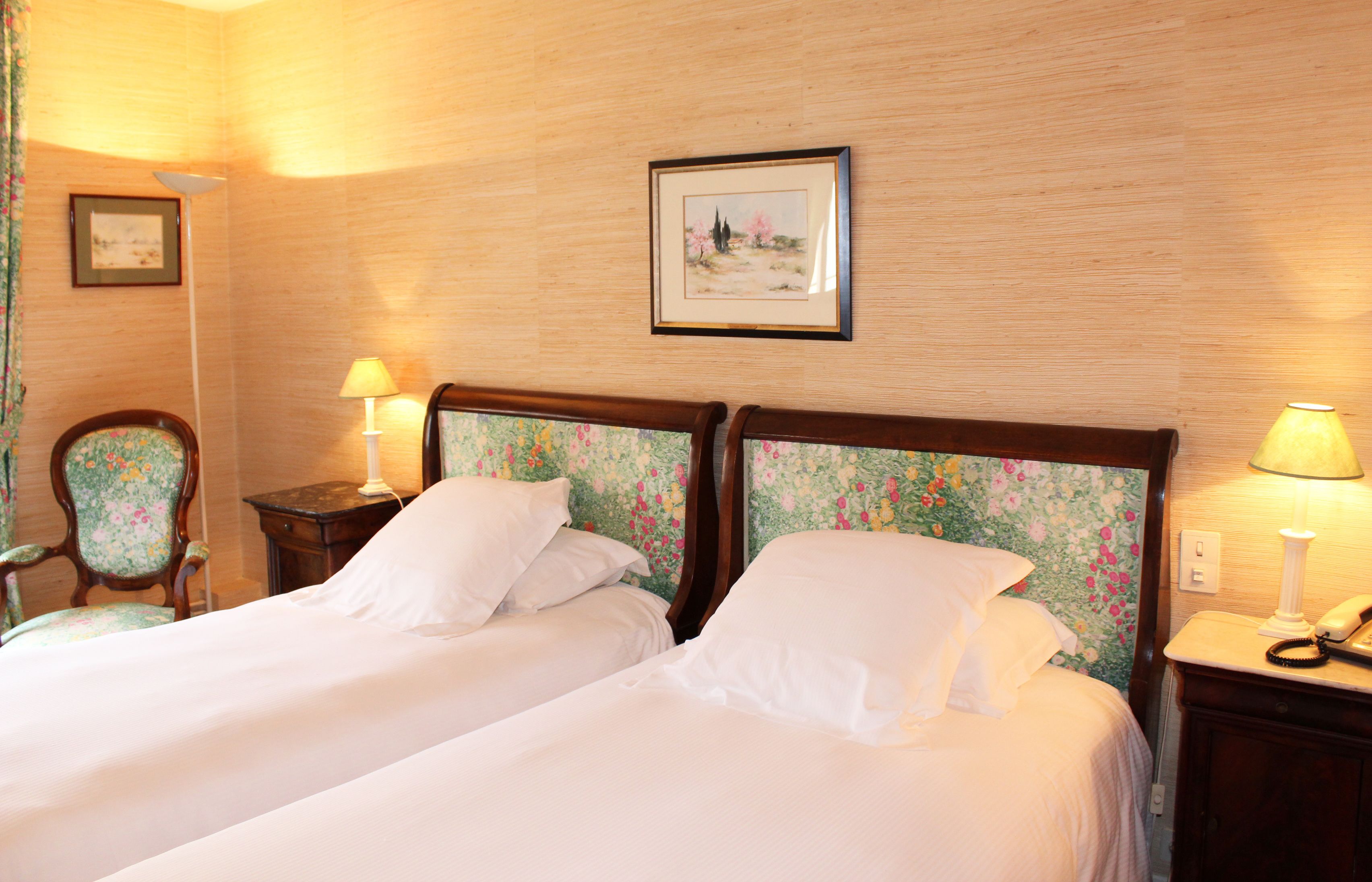 Une chambre d’hôtel avec deux lits, des têtes de lit à motifs floraux, des lampes de chevet, une chaise et un tableau encadré au mur.