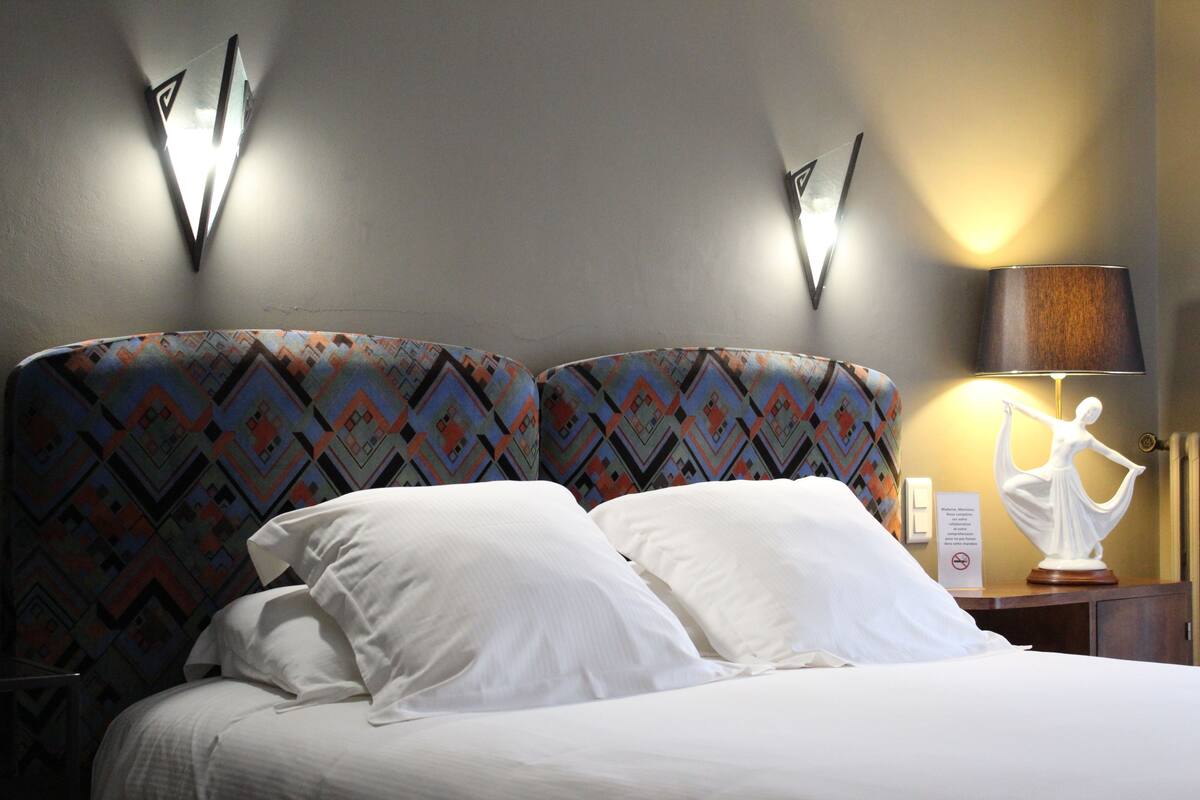 A hotel room with a bed, patterned headboards, a nightstand with a lamp, and a wall-mounted light fixture.