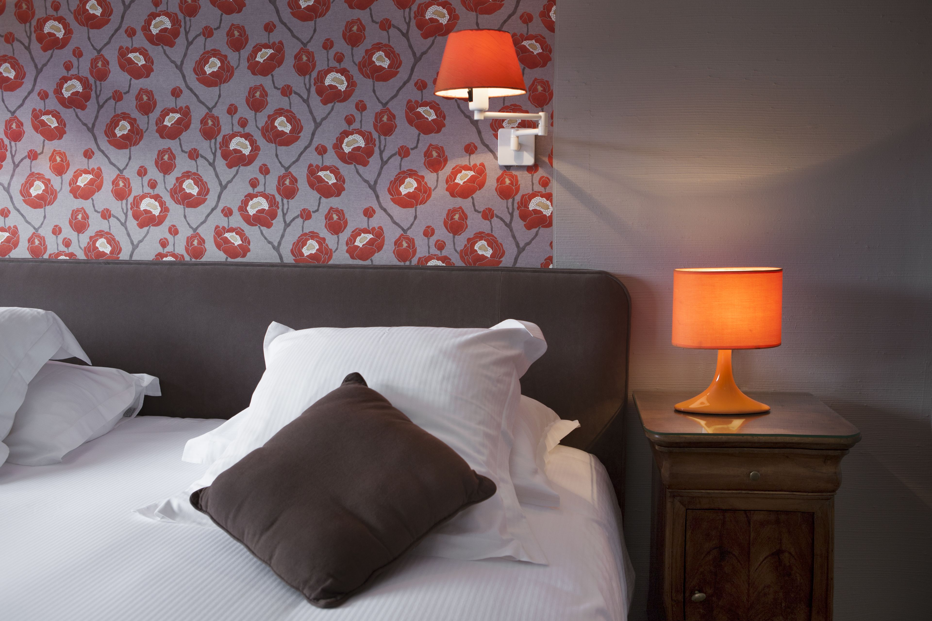 A neatly made bed with white and brown pillows, a red floral wallpaper, and a wooden nightstand with a lamp.