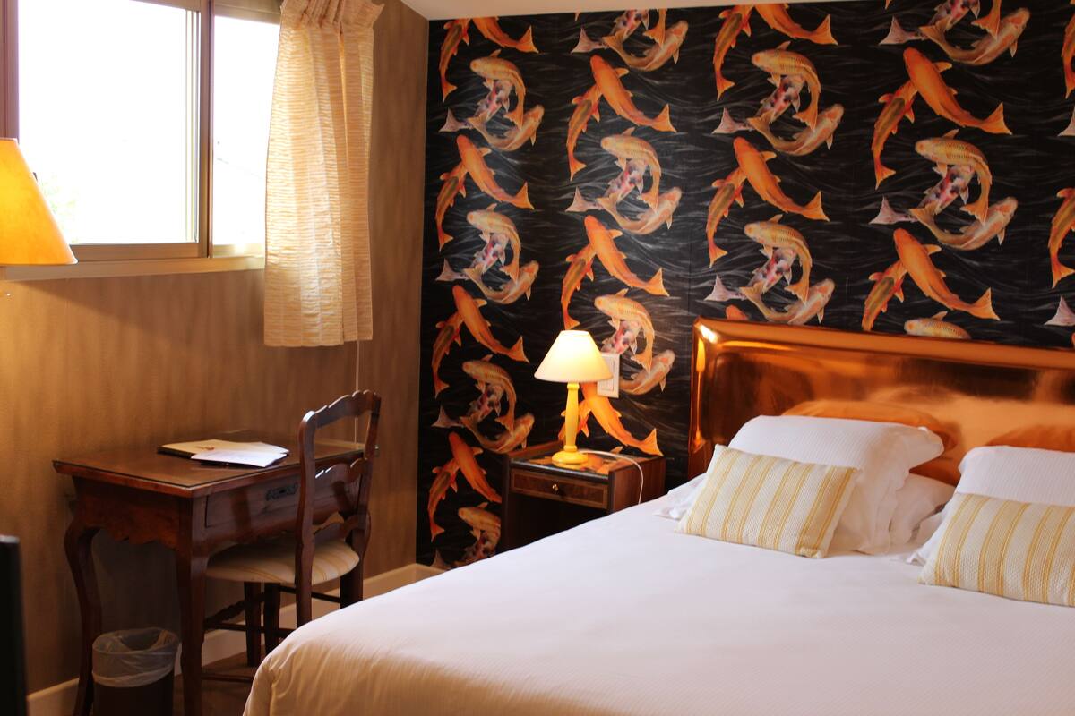A hotel room with a bed, a desk, a chair, a lamp, and a wall with a fish pattern.