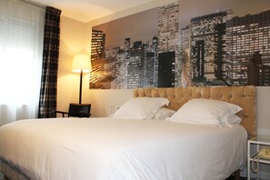 Habitación de hotel con una cama grande, un mural de paisaje urbano en la pared, una lámpara y un teléfono en una mesita de noche.