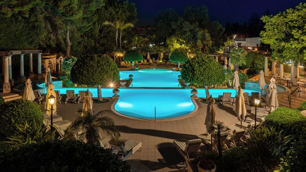 A nighttime view of a resort's outdoor pool area with two pools, surrounded by lounge chairs, umbrellas, and well-maintained landscaping. - Corinthia Palace Malta (Attard)