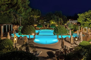 A nighttime view of a resort's outdoor pool area with two pools, surrounded by lounge chairs, umbrellas, and well-maintained landscaping. - Corinthia Palace Malta (Attard)