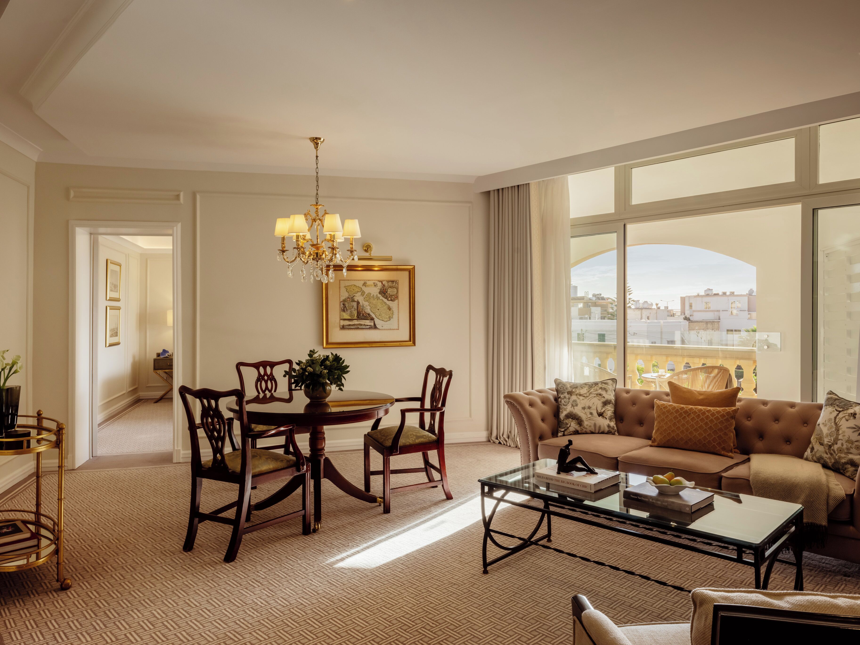 A spacious living room with a large window, a chandelier, a dining area with a round table and chairs, and a sofa with decorative pillows.