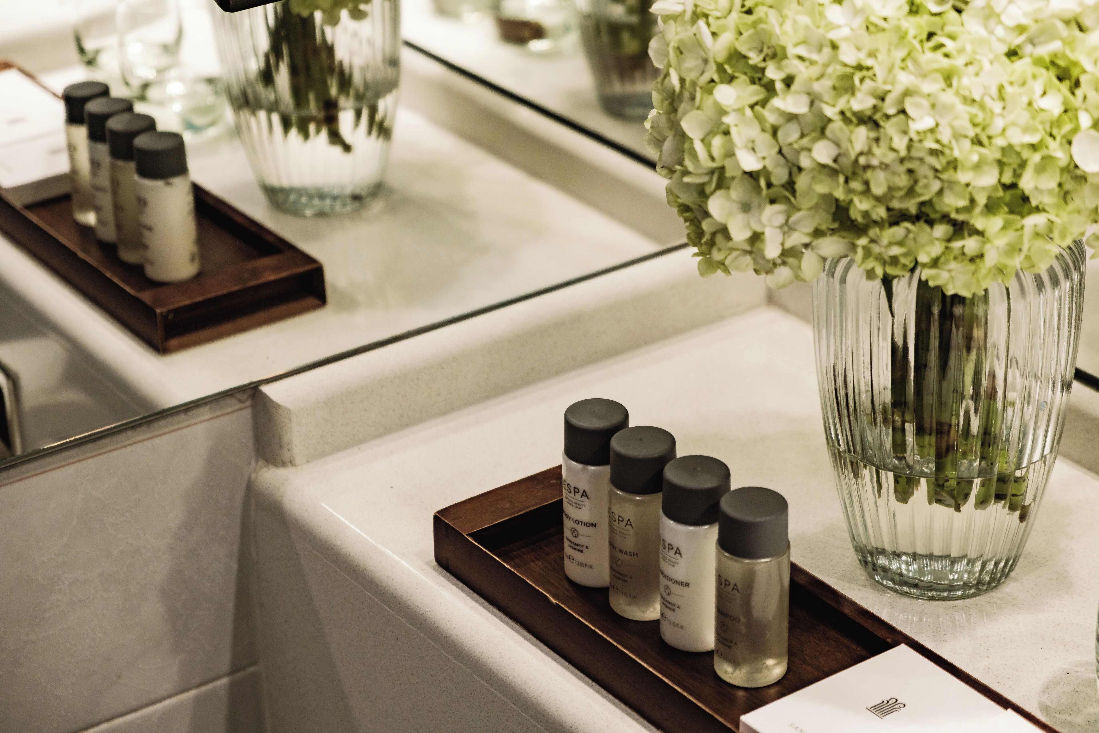 Una encimera de baño con una bandeja que contiene varias botellas de productos de spa, un florero con flores blancas y un espejo que refleja la escena.
