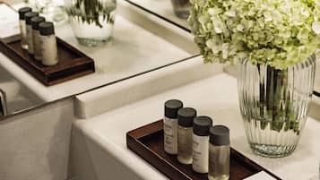 Una encimera de baño con una bandeja que contiene varias botellas de productos de spa, un florero con flores blancas y un espejo que refleja la escena.
