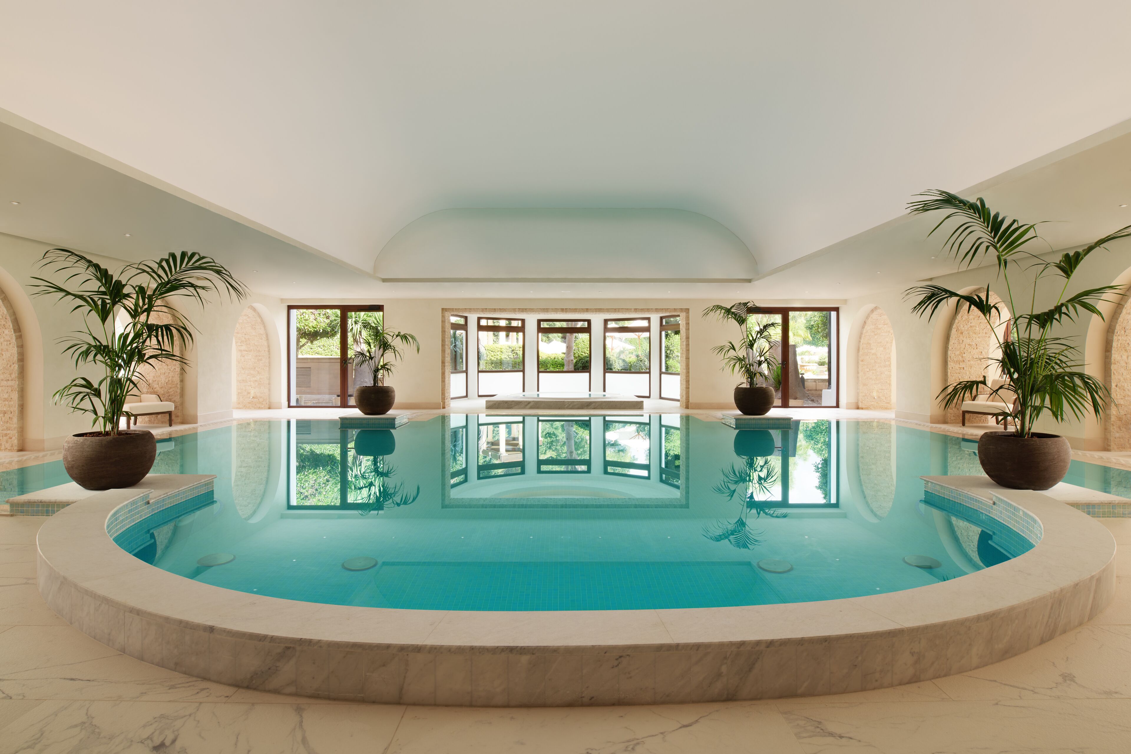 Una amplia piscina interior con vistas despejadas al exterior gracias a grandes ventanales, rodeada de plantas en macetas.