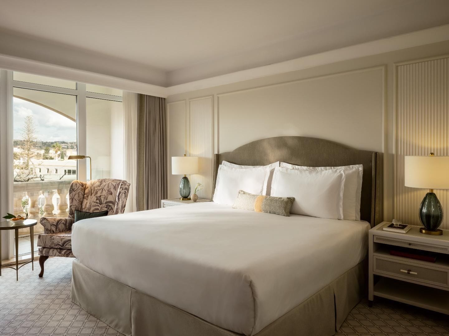 A large bed with white linens, two bedside tables with lamps, a chair, and a view of the city through large windows.