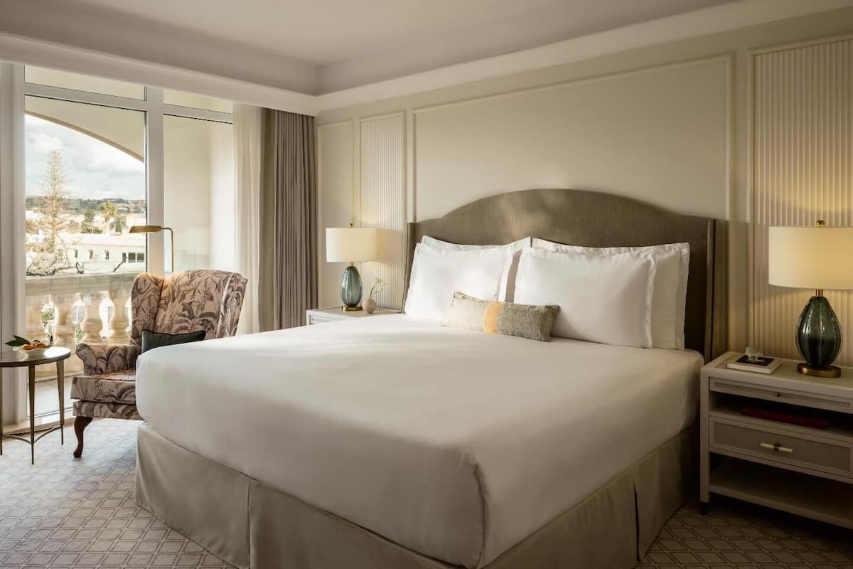 A large bed with white linens, two bedside tables with lamps, a chair, and a view of the city through large windows.