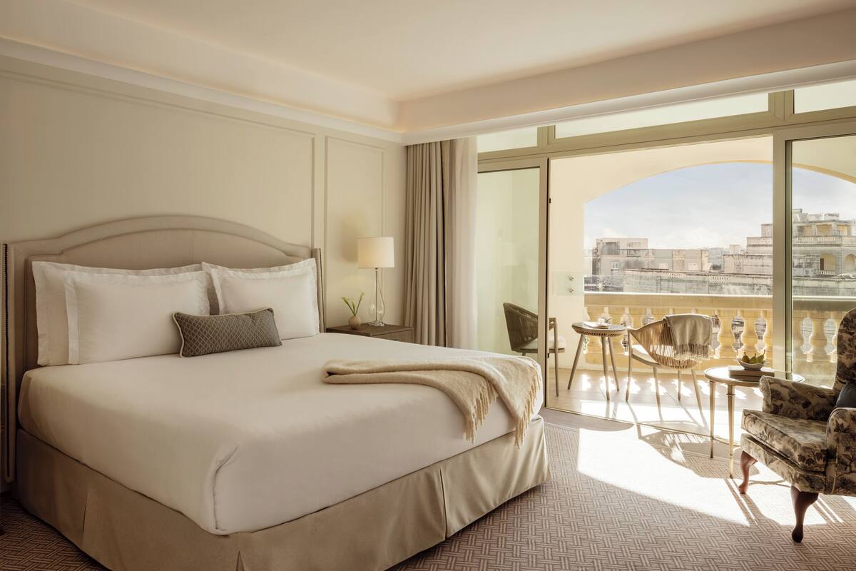 A hotel room with a large bed, a balcony with a view of the city, and a seating area with a table and chairs.