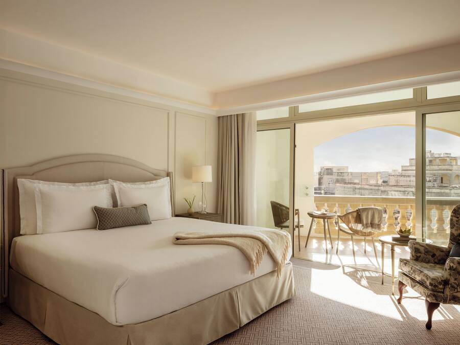 Habitación de hotel con una cama grande, un balcón con vistas a la ciudad y un espacio de descanso con una mesa y sillas.