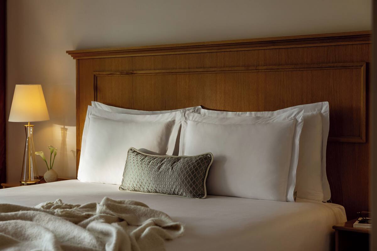 A neatly made bed with white linens and a textured pillow, a bedside lamp, and a vase with flowers on a wooden nightstand.