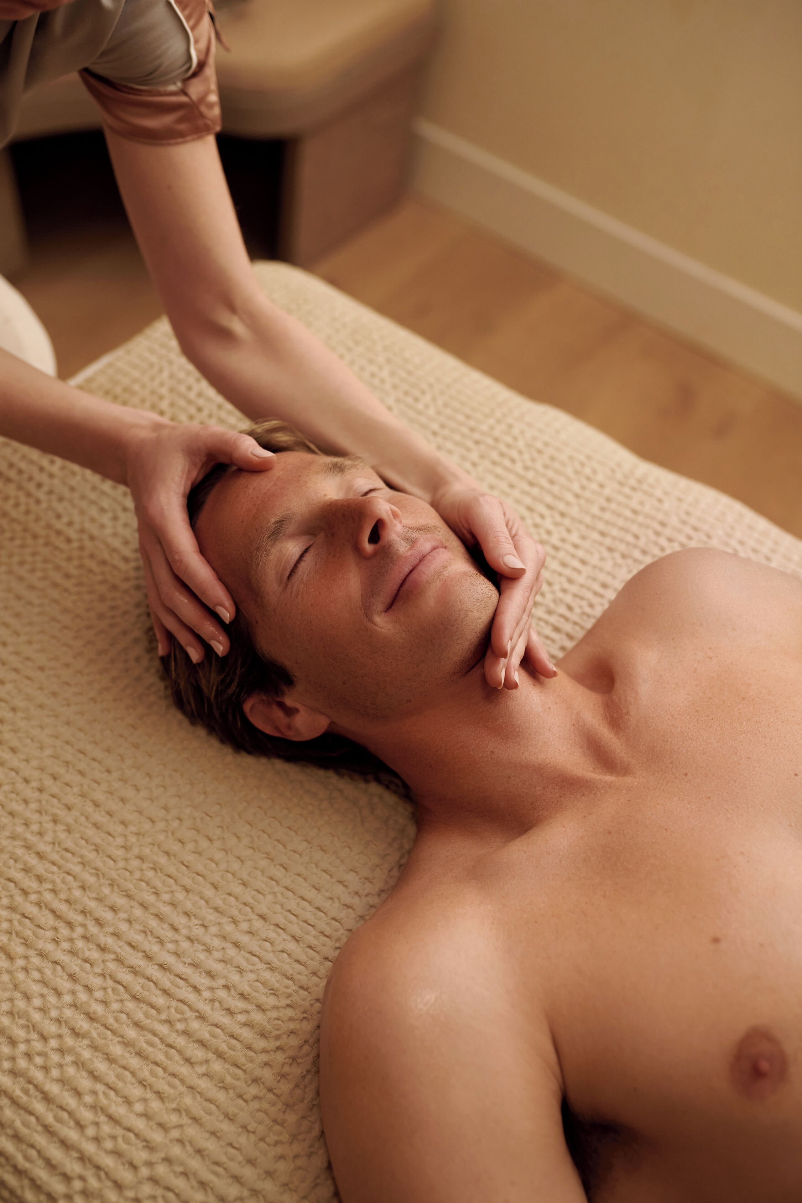 A person receiving a head massage on a textured beige mat in a room with wooden flooring and a neutral-colored wall.