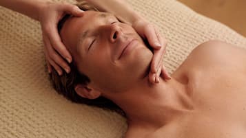 A person receiving a head massage on a textured beige mat in a room with wooden flooring and a neutral-colored wall.