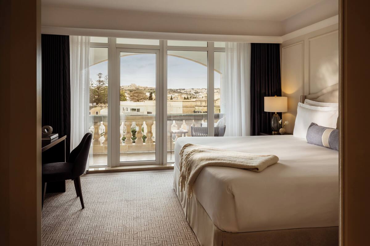 A hotel room with a large bed, a desk, and a view of the city through floor-to-ceiling windows.