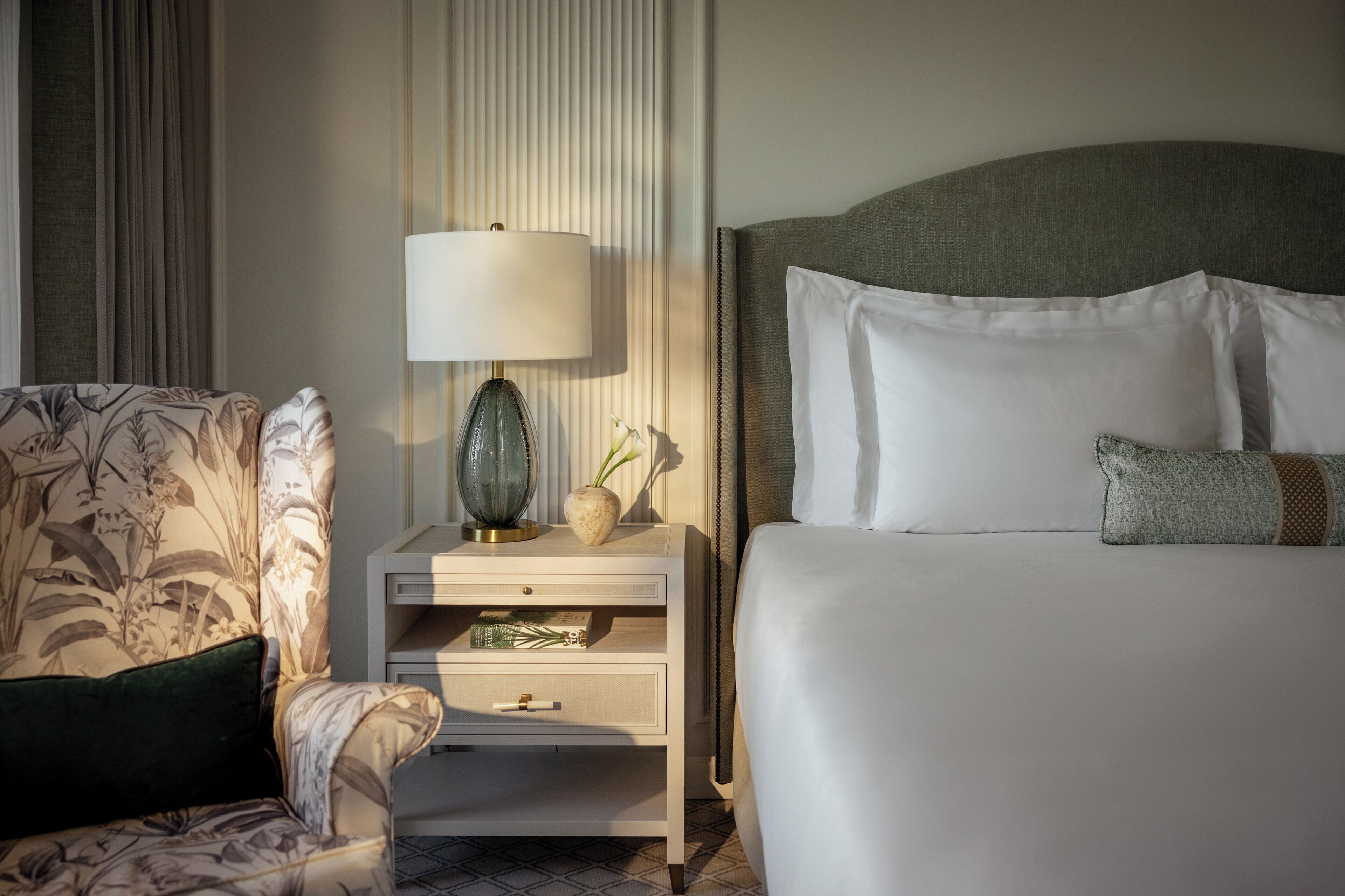 A hotel room with a bed, a nightstand with a lamp, a chair, and a decorative vase.