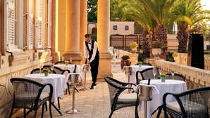 An outdoor dining area with tables and chairs, a waiter, and palm trees. - Corinthia Palace Malta (Attard)
