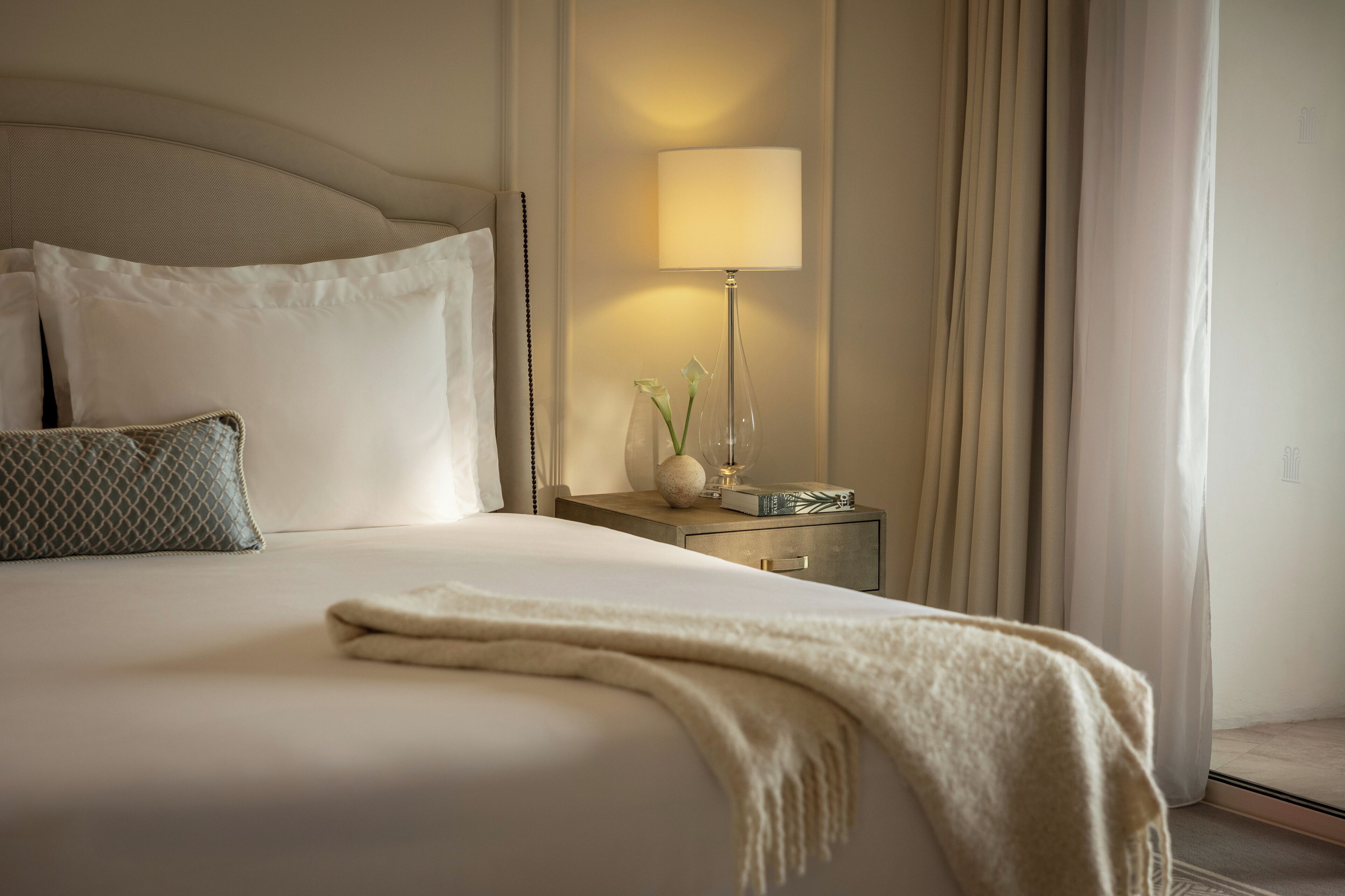 Una cama bien hecha con edredón blanco y manta color beige, una mesita de noche con lámpara, y un florero con flores.
