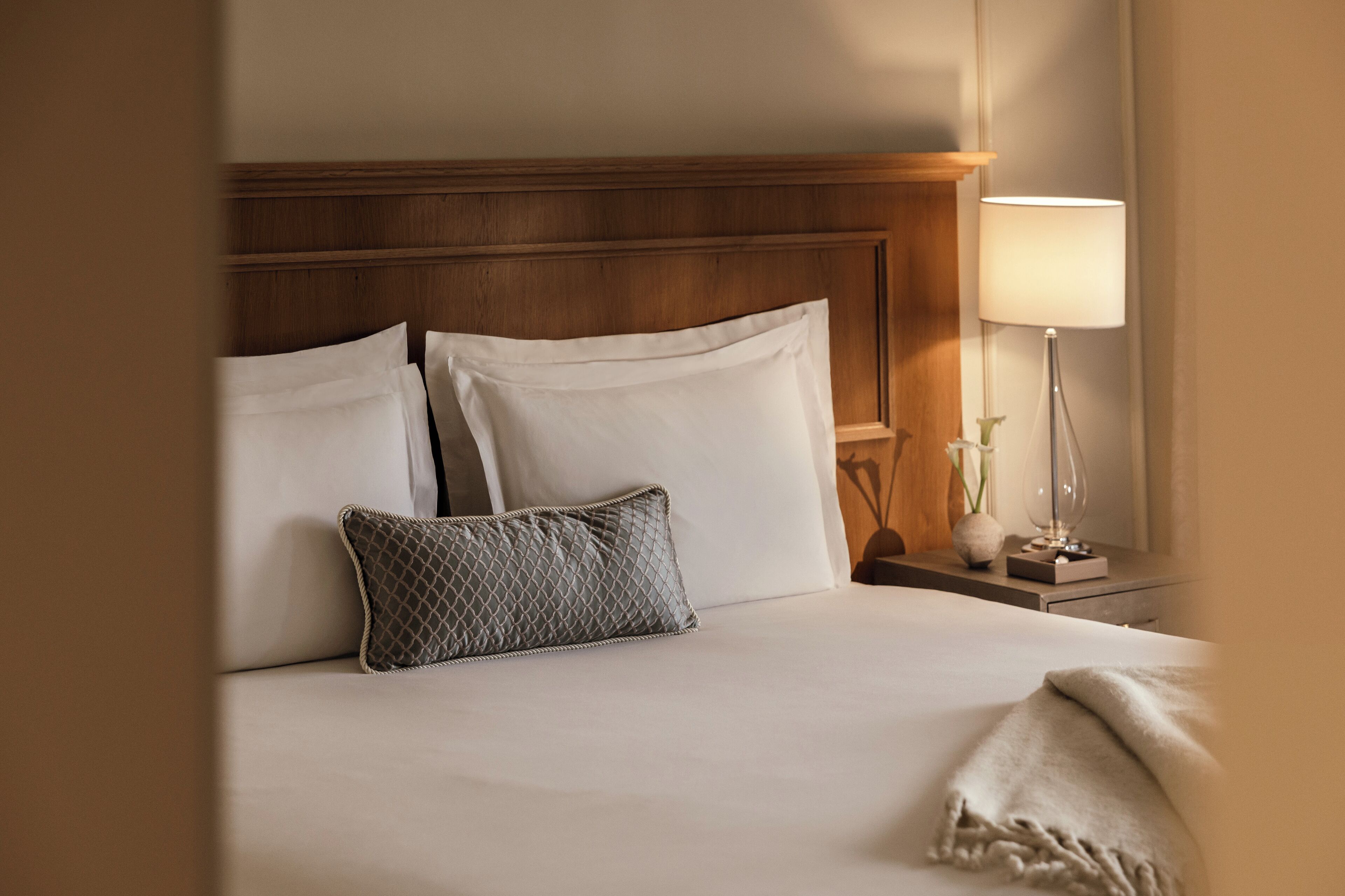 A neatly made bed with white linens and a grey patterned pillow, a wooden headboard, and a bedside table with a lamp and a vase of flowers.