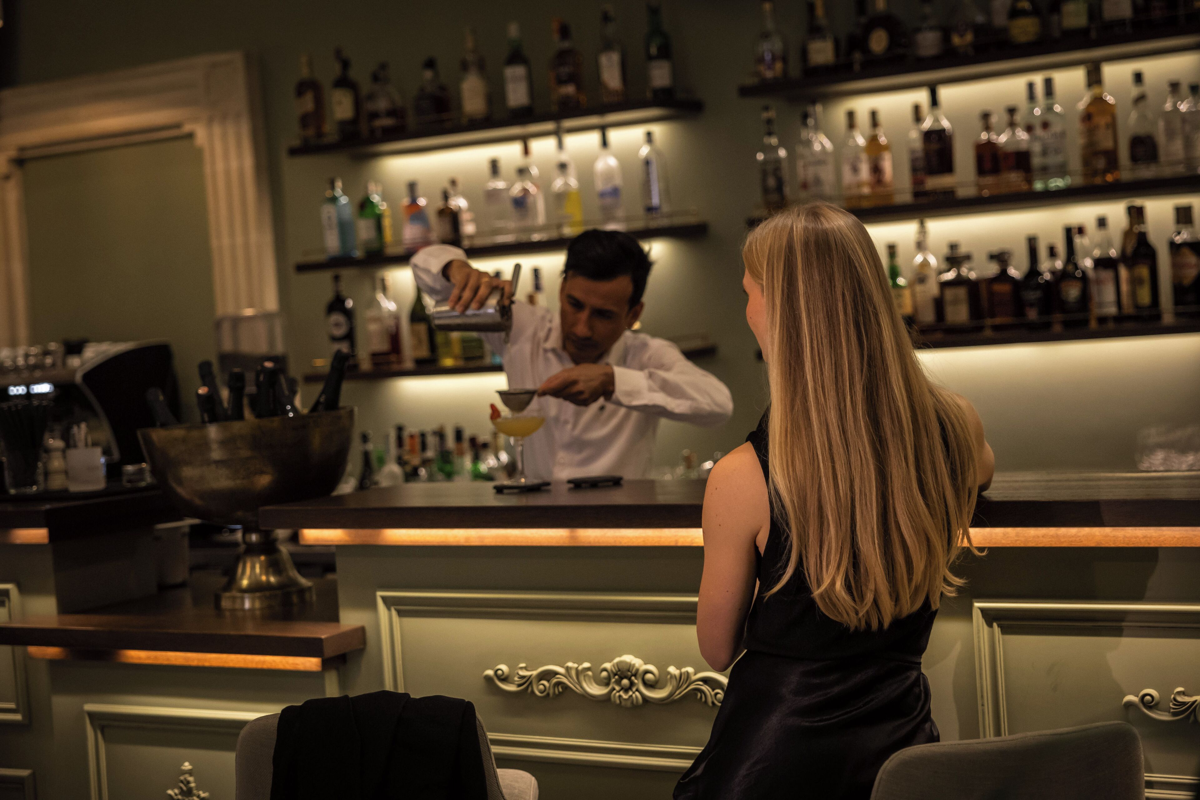 Zona de bar con un cantinero preparando una bebida, estantes con botellas de licor y una barra.