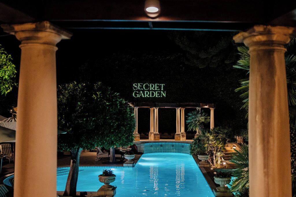 Een nachtzicht van een zwembadgedeelte met kolommen en een bord waarop staat 'SECRET GARDEN'.