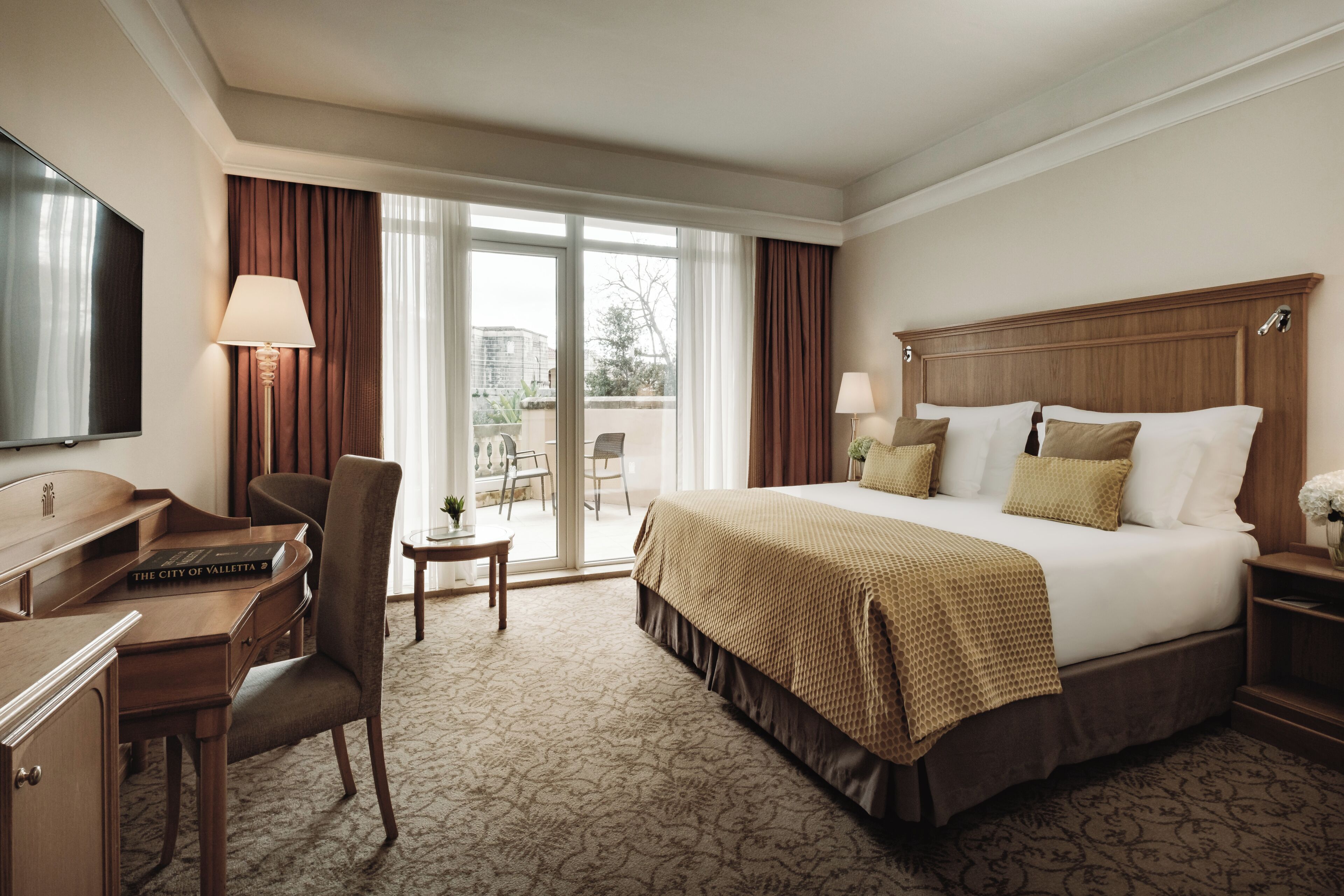 A hotel room with a large bed, a desk with a computer, a chair, a TV, and a view of the outdoors through a sliding door.